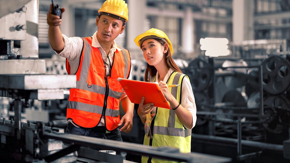 Baret ve reflektörlü yelek giyen iki iş güvenliği uzmanı fabrikada denetim yapıyor